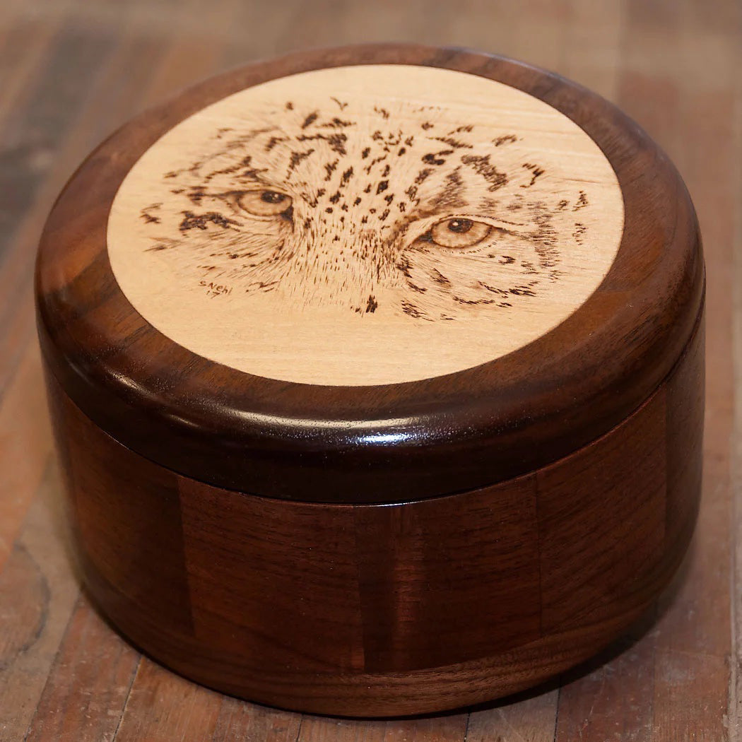 A woodturned round, wooden, lidded box. The box is made of walnut and the lid has a light colored inset with a Cheetah's eyes woodburned on the top. 