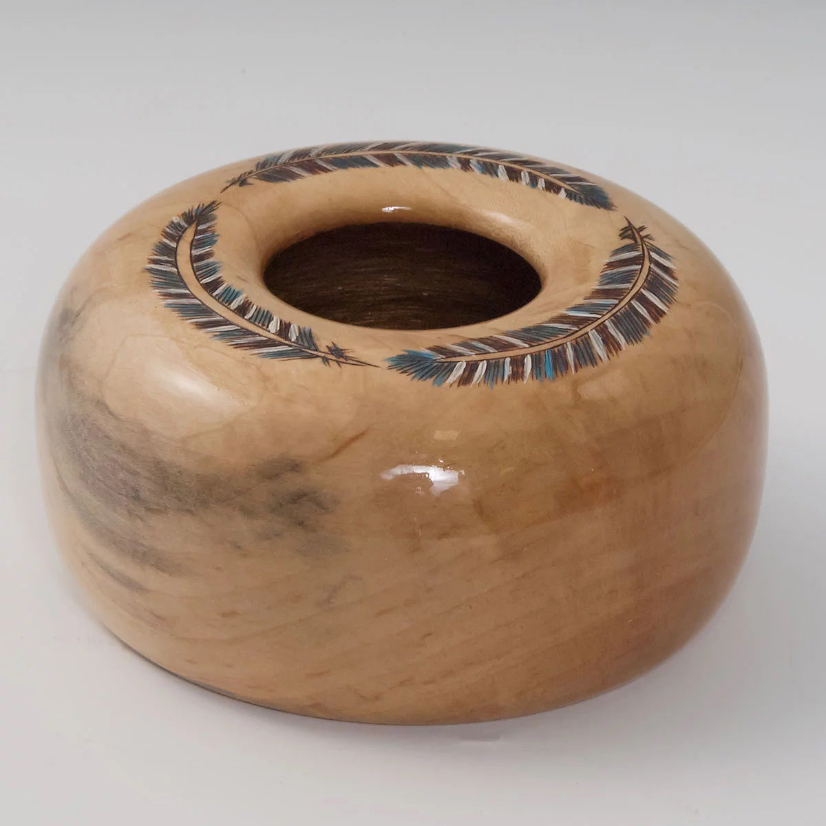 A light-colored woodturned vessel with feathers burned on the top edge. 