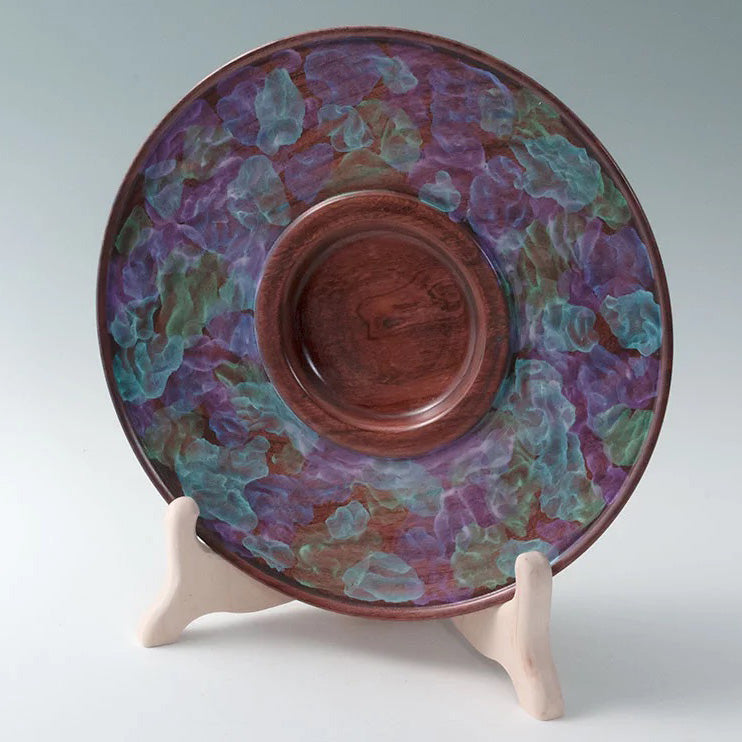 A woodturned platter on a stand. The platter is painted with blue, green and purple leaves with a cherry-colored wooden center. 