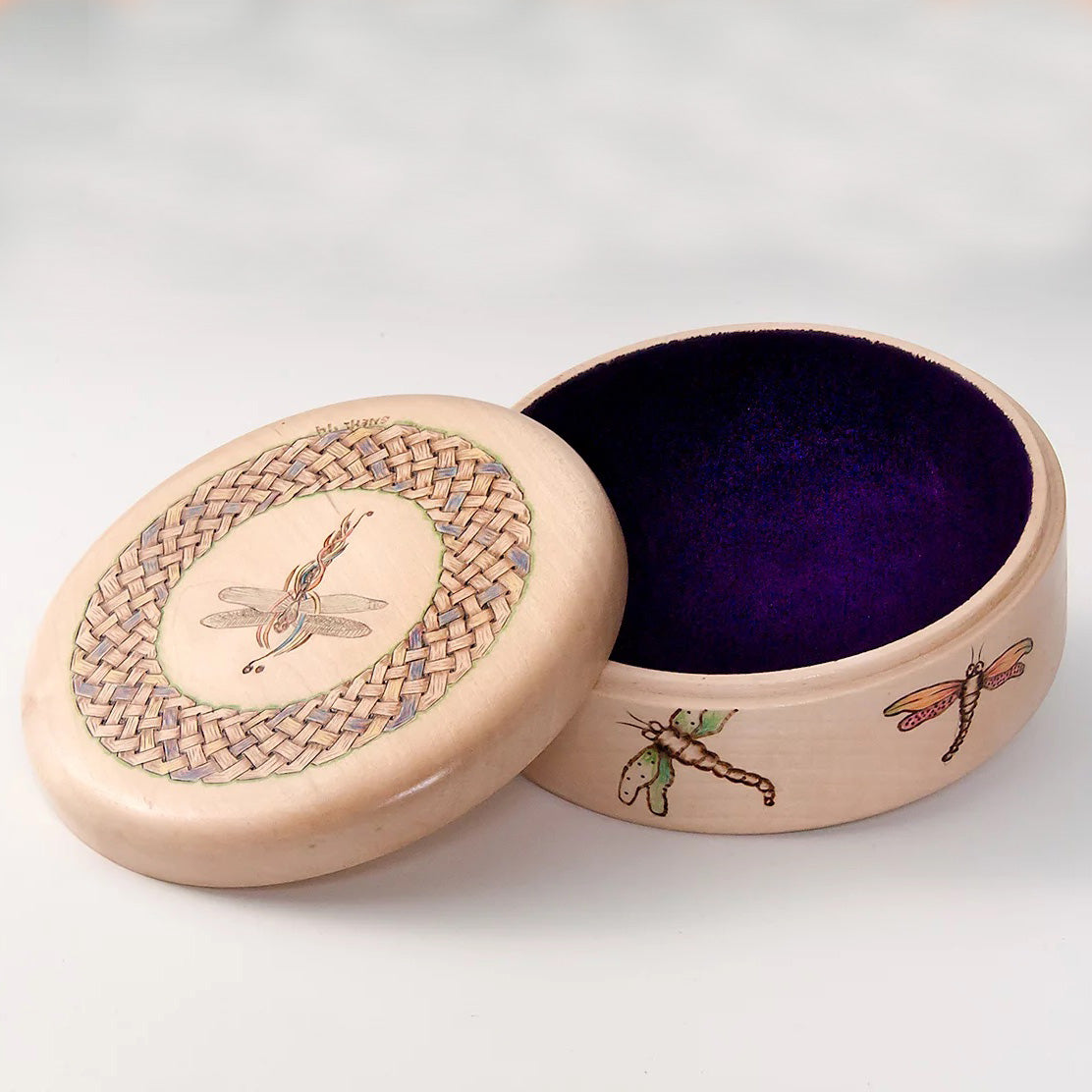 A round, lidded box with a felt bottom. The box is adorned with woodburned and hand-painted dragonflies. 